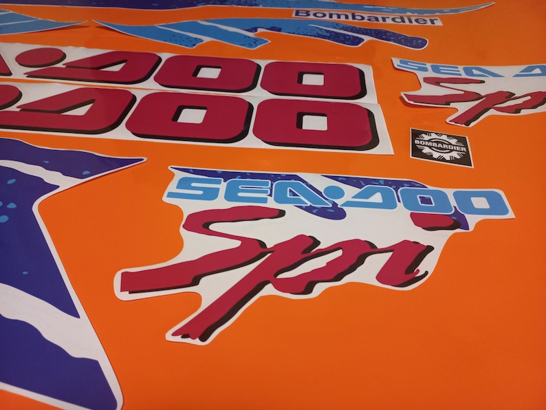 K&ouml;nnte beinhalten: Ein Satz roter und blauer Sea-Doo-Aufkleber mit dem Text "Sea-Doo" und "Sea-Doo Sport" sowie der Nummer "2000". Die Aufkleber befinden sich auf einem leuchtend orangenen Hintergrund.
