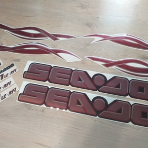 May include: A set of red and white Sea-Doo decals. The decals include two large Sea-Doo logos, three smaller Sea-Doo logos, and four red and white flame-shaped decals. The decals are designed for a Sea-Doo GTI LE-RFi.