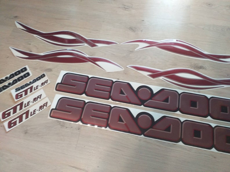 May include: A set of red and white Sea-Doo decals. The decals include two large Sea-Doo logos, three smaller Sea-Doo logos, and four red and white flame-shaped decals. The decals are designed for a Sea-Doo GTI LE-RFi.