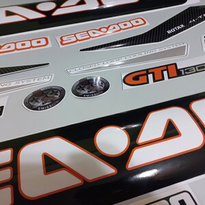 May include: A set of black and white Sea-Doo decals with orange accents. The decals include the words "Sea-Doo", "GTI 130", "Closed-Loop Cooling System", "Rotax 4-TEC", and "Intelligent Brake System".