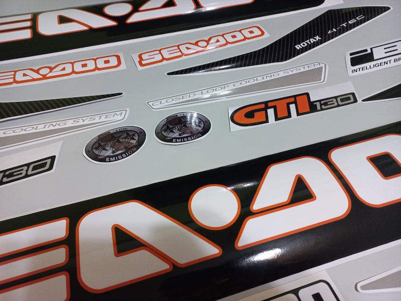 May include: A set of black and white Sea-Doo decals with orange accents. The decals include the words "Sea-Doo", "GTI 130", "Closed-Loop Cooling System", "Rotax 4-TEC", and "Intelligent Brake System".