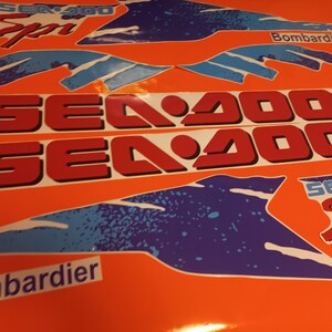 K&ouml;nnte beinhalten: Ein Satz rot-wei&szlig;er Sea-Doo-Aufkleber mit blauen und wei&szlig;en Akzenten. Die Aufkleber zeigen den Text "Sea-Doo" und "Sea-Doo Sport" sowie das Logo "Bombardier".