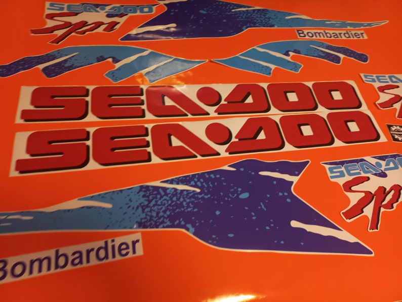 K&ouml;nnte beinhalten: Ein Satz rot-wei&szlig;er Sea-Doo-Aufkleber mit blauen und wei&szlig;en Akzenten. Die Aufkleber zeigen den Text "Sea-Doo" und "Sea-Doo Sport" sowie das Logo "Bombardier".