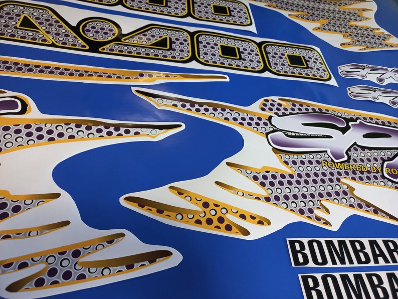 Puede incluir: Un conjunto de calcoman&iacute;as blancas y doradas con un dise&ntilde;o estampado en blanco y negro. Las calcoman&iacute;as presentan el texto "SP" y "POWERED BY ROX" y un patr&oacute;n geom&eacute;trico. Las calcoman&iacute;as est&aacute;n dise&ntilde;adas para un fondo azul.