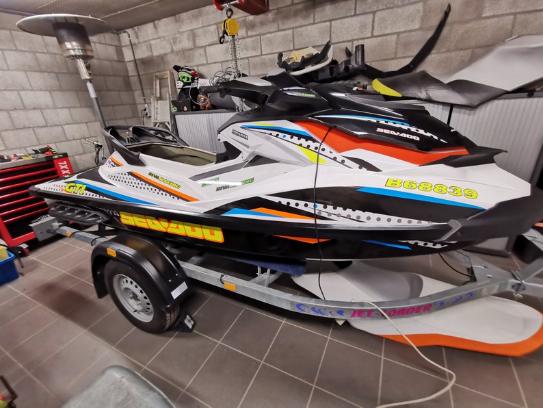 May include: A white, black, orange and blue Sea-Doo Spark jet ski with a black and white graphic design. The jet ski is on a trailer with a black tyre and the registration number B68839.