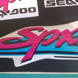Puede incluir: Una pegatina "Sea-Doo" rosa y negra con un dise&ntilde;o de rayas azules y blancas. La pegatina dice "Sea-Doo" y "Spa" en una fuente estilizada.