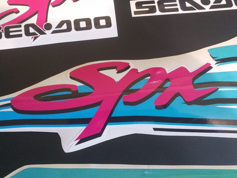 Puede incluir: Una pegatina "Sea-Doo" rosa y negra con un dise&ntilde;o de rayas azules y blancas. La pegatina dice "Sea-Doo" y "Spa" en una fuente estilizada.