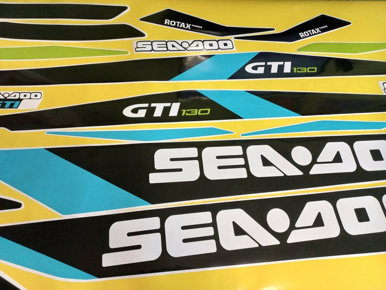 以下が含まれることがあります： 青と緑のアクセントが付いた白黒のシー-ドゥーデカール。デカールには「シー-ドゥー」、「GTI 130」、「ROTAX」の文字が書かれています。