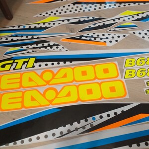 May include: A set of black, white, blue, orange and yellow decals with the text "Sea Doo GTI" and "B6883" on a white background. The decals are for a Sea Doo watercraft.