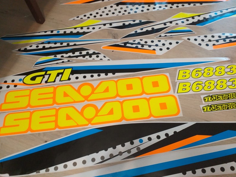 May include: A set of black, white, blue, orange and yellow decals with the text "Sea Doo GTI" and "B6883" on a white background. The decals are for a Sea Doo watercraft.