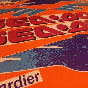 K&ouml;nnte beinhalten: Ein Satz pinkfarbener und schwarzer Sea-Doo-Aufkleber mit einem blauen und violetten Grafikdesign. Die Aufkleber befinden sich auf einem orangefarbenen Hintergrund.