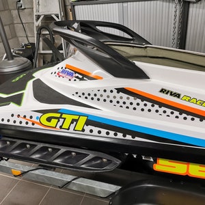 May include: A white and black jet ski with a yellow and orange "RIVA RACING" logo on the side. The jet ski has a black and white polka dot pattern and a yellow "GTI" logo.