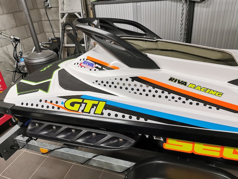 May include: A white and black jet ski with a yellow and orange "RIVA RACING" logo on the side. The jet ski has a black and white polka dot pattern and a yellow "GTI" logo.