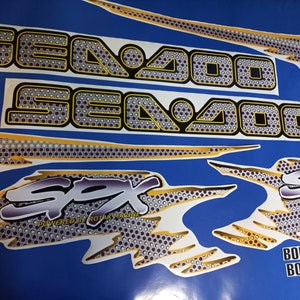 Puede incluir: Un conjunto de calcoman&iacute;as Sea-Doo blancas y doradas con un patr&oacute;n de panal de abeja blanco y negro. Las calcoman&iacute;as presentan el texto "Sea-Doo", "SPX", "Powered by Rotax Marine" y "Bombardier".