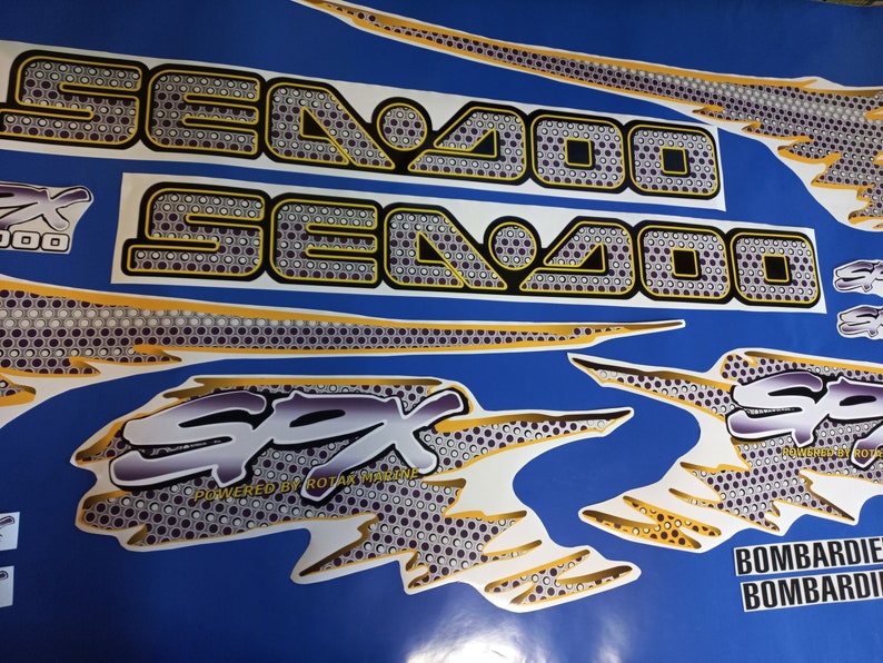 Puede incluir: Un conjunto de calcoman&iacute;as Sea-Doo blancas y doradas con un patr&oacute;n de panal de abeja blanco y negro. Las calcoman&iacute;as presentan el texto "Sea-Doo", "SPX", "Powered by Rotax Marine" y "Bombardier".