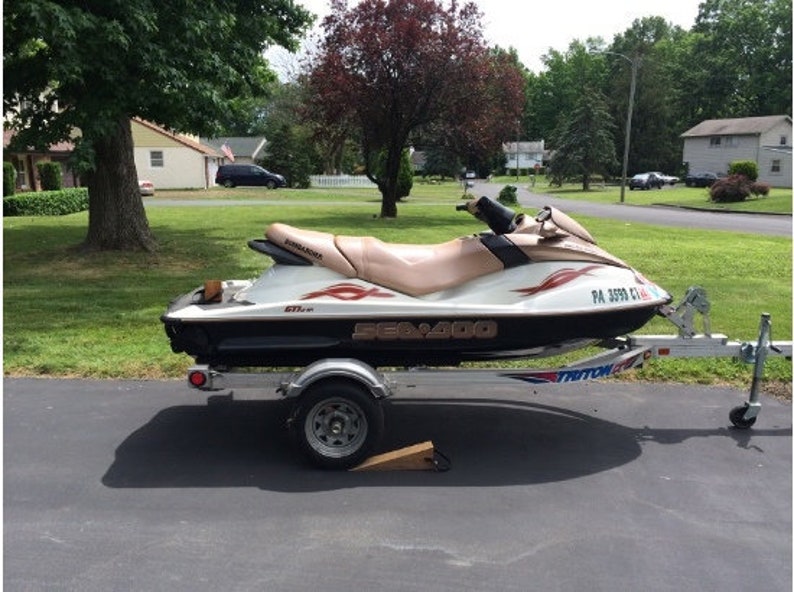 May include: A white and black Sea-Doo 650cc jet ski on a trailer with a black and white Triton logo. The jet ski has a tan seat and red and white graphics. The trailer has a black wheel and a black and white license plate that reads "PA 3593 CX".