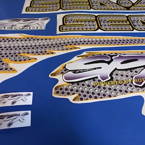 Puede incluir: Un conjunto de calcoman&iacute;as blancas y moradas con detalles dorados. Las calcoman&iacute;as presentan las palabras "Sea-Doo" y "SPX" con un patr&oacute;n de panal. Las calcoman&iacute;as est&aacute;n dise&ntilde;adas para una moto acu&aacute;tica Sea-Doo.