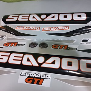 May include: A set of black and white Sea-Doo decals with orange accents. The decals feature the Sea-Doo logo, the text "Sea-Doo", "Sea-Doo GTI", "GTI 130", "Intelligent Brake & Reverse", and "Closed-Loop Cooling System".