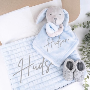 May include: A baby gift set in a box, featuring a light blue plush blanket and a matching security blanket with the name "Hudson" in script. Includes a gray bunny plush toy and a pair of gray knit baby booties.