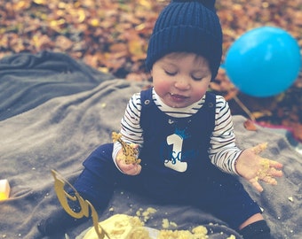 May include: A baby wearing a blue and white striped shirt and a blue knitted hat with a pom-pom sits on a gray blanket. The baby is holding a toy giraffe and has a cake in front of them. The cake is decorated with a number one and a giraffe.