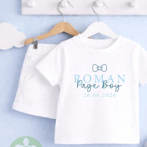 May include: A white baby outfit set, comprising a t-shirt and shorts, displayed on a wooden hanger. The t-shirt has the text "ROMAN Page Boy" in blue, a bow tie graphic, and the date 26.06.2026.