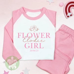 May include: A pink and white pyjama set. The top has pink sleeves and a white body with the words "FLOWER Elodie GIRL" printed on the front. The matching trousers are solid pink. The date "20/06/25" is printed below the text.