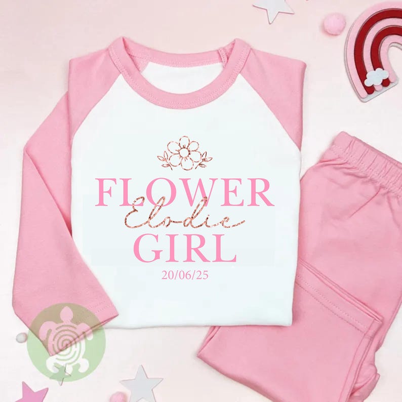 May include: A pink and white pyjama set. The top has pink sleeves and a white body with the words "FLOWER Elodie GIRL" printed on the front. The matching trousers are solid pink. The date "20/06/25" is printed below the text.