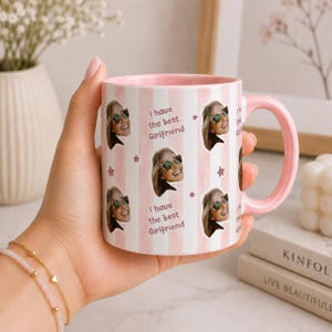 May include: A ceramic mug with a light pink and white vertical striped pattern. The mug features repeating photo cutouts of a person wearing sunglasses, small pink stars, and the printed text "I have the best Girlfriend" in a dark pink font.