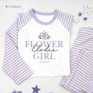 May include: A lavender and white striped pyjama set. The top has lavender sleeves and a white front with the text "FLOWER Elodie GIRL" and a flower graphic. The matching trousers are lavender and white striped. The date "20/06/25" is printed on the shirt.