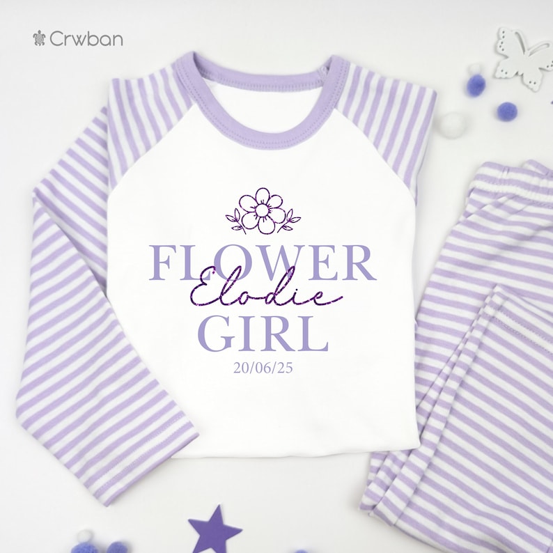 May include: A lavender and white striped pyjama set. The top has lavender sleeves and a white front with the text "FLOWER Elodie GIRL" and a flower graphic. The matching trousers are lavender and white striped. The date "20/06/25" is printed on the shirt.