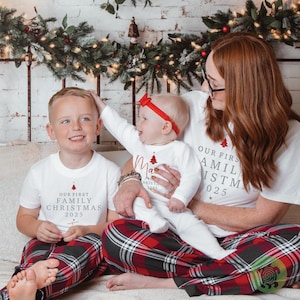 May include: White t-shirts and a long-sleeved onesie featuring the red text Our First Family Christmas 2025 with a small red tree graphic. The clothing is paired with red, black, and white plaid pajama pants, and the infant wears a red bow headband.
