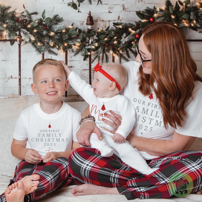 May include: White t-shirts and a long-sleeved onesie featuring the red text Our First Family Christmas 2025 with a small red tree graphic. The clothing is paired with red, black, and white plaid pajama pants, and the infant wears a red bow headband.