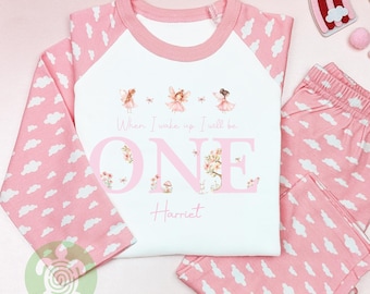 May include: A pink and white pajama set for a child. The long-sleeved top has pink sleeves with a cloud pattern and a white front with the text "When I wake up I will be ONE Harriet" and fairy illustrations. The matching pants have a pink cloud pattern.