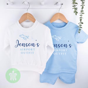 May include: Two white and blue airplane themed baby outfits. One is a long-sleeved shirt with the text "Jenson's Airport Outfit" and the other is a short-sleeved shirt with the text "Jenson's Airport Outfit".