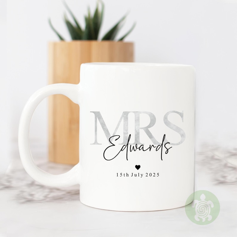 May include: White ceramic mug with the text "MRS Edwards" in silver and black script. Below the name is "15th July 2025" and a small heart. A wooden planter with a green plant is in the background.