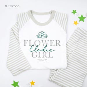May include: A white and grey striped pyjama set featuring the text "FLOWER Elodie GIRL" and the date "20/06/25". The long-sleeved top has a white body and grey striped sleeves. A green flower graphic is above the text.