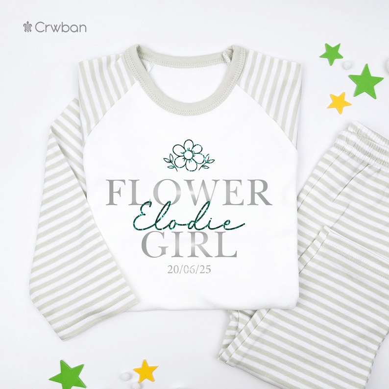 May include: A white and grey striped pyjama set featuring the text "FLOWER Elodie GIRL" and the date "20/06/25". The long-sleeved top has a white body and grey striped sleeves. A green flower graphic is above the text.