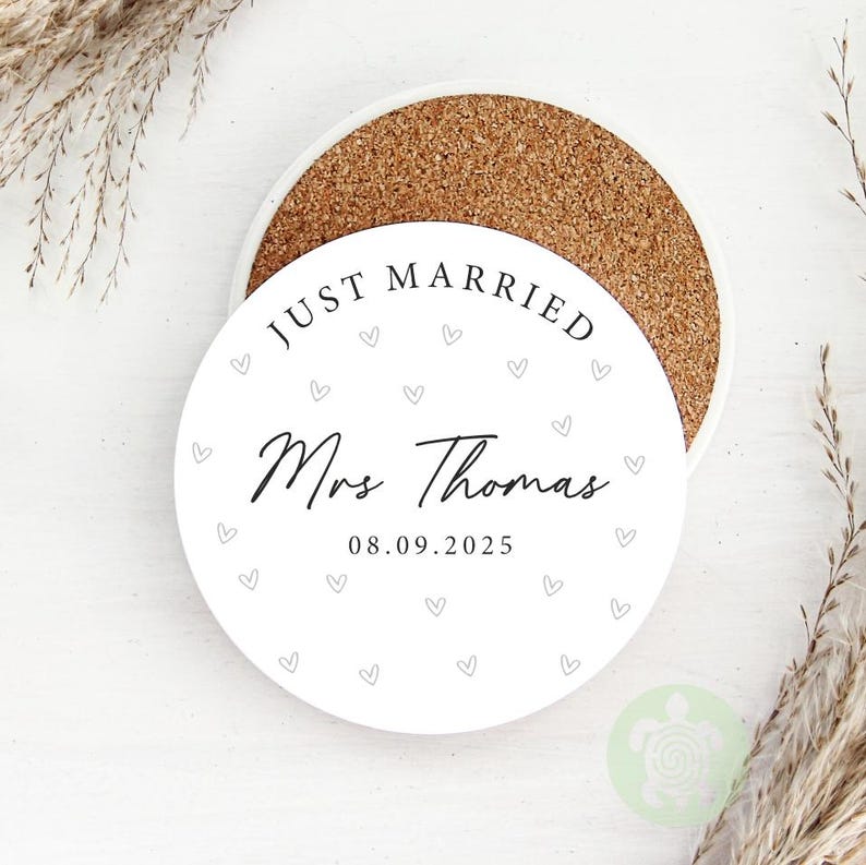 May include: A round white coaster with the text "JUST MARRIED Mrs Thomas 08.09.2025" and small gray hearts. The coaster sits on a cork base. Decorative dried plants are in the background.