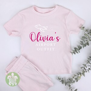 May include: Pink short-sleeved t-shirt with a white airplane graphic and the text "Olivia's Airport Outfit" in pink.