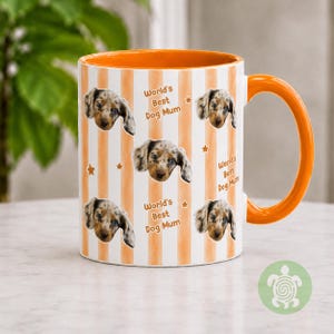 May include: A ceramic mug with an orange interior and handle, featuring a white and orange vertical striped pattern. The exterior is decorated with repeated images of a merle dachshund puppy face, small orange stars, and the text World's Best Dog Mum.