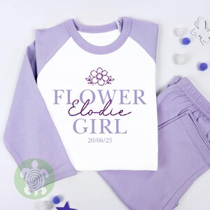 May include: A lavender and white pyjama set featuring the text "FLOWER Elodie GIRL" and a floral design. The top has lavender sleeves and a white body. The matching trousers are lavender.