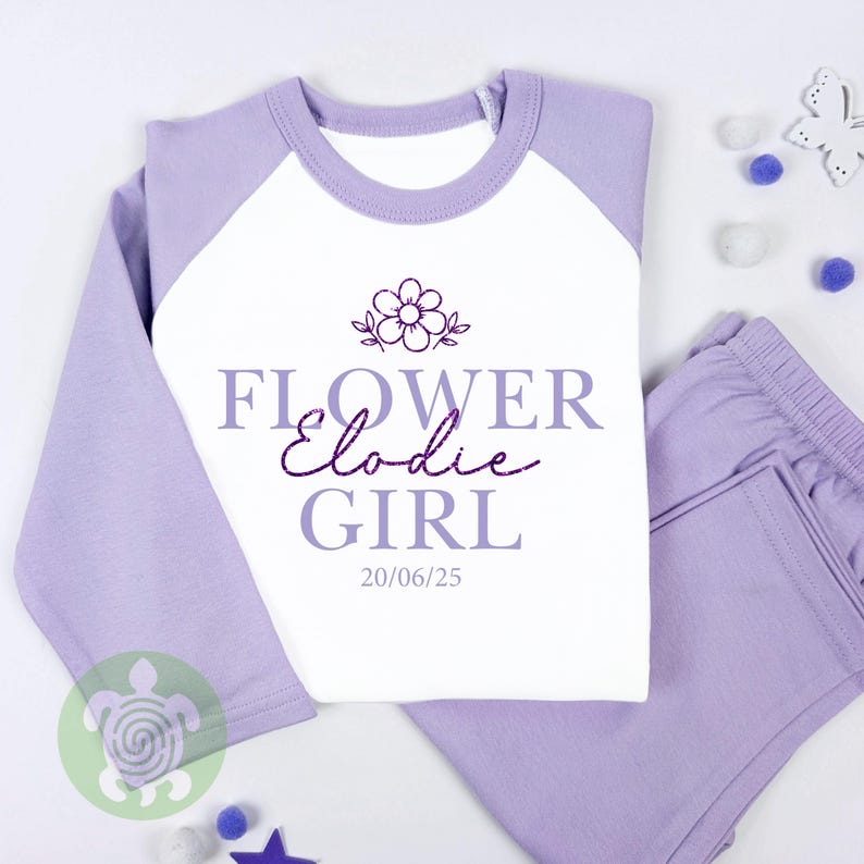 May include: A lavender and white pyjama set featuring the text "FLOWER Elodie GIRL" and a floral design. The top has lavender sleeves and a white body. The matching trousers are lavender.