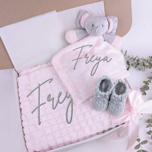 Puede incluir: Un juego de regalo para beb&eacute; en una caja, con una manta rosa y una manta de seguridad a juego, ambas bordadas con el nombre "Freya". Incluye un peluche de elefante gris y un par de patucos grises. La caja est&aacute; atada con una cinta rosa.