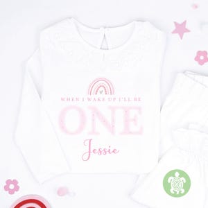 May include: White long-sleeved top with a lace collar, featuring the text "WHEN I WAKE UP I'LL BE ONE Jessie" in pink, and a rainbow graphic. White trousers are also visible.
