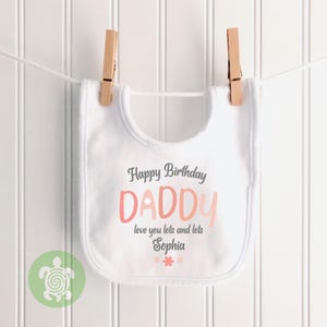 May include: White baby bib with the text "Happy Birthday DADDY" in gray and pink. The bib is hanging from a clothesline with wooden clothespins. The text "love you lots and lots Sophia" is also on the bib.