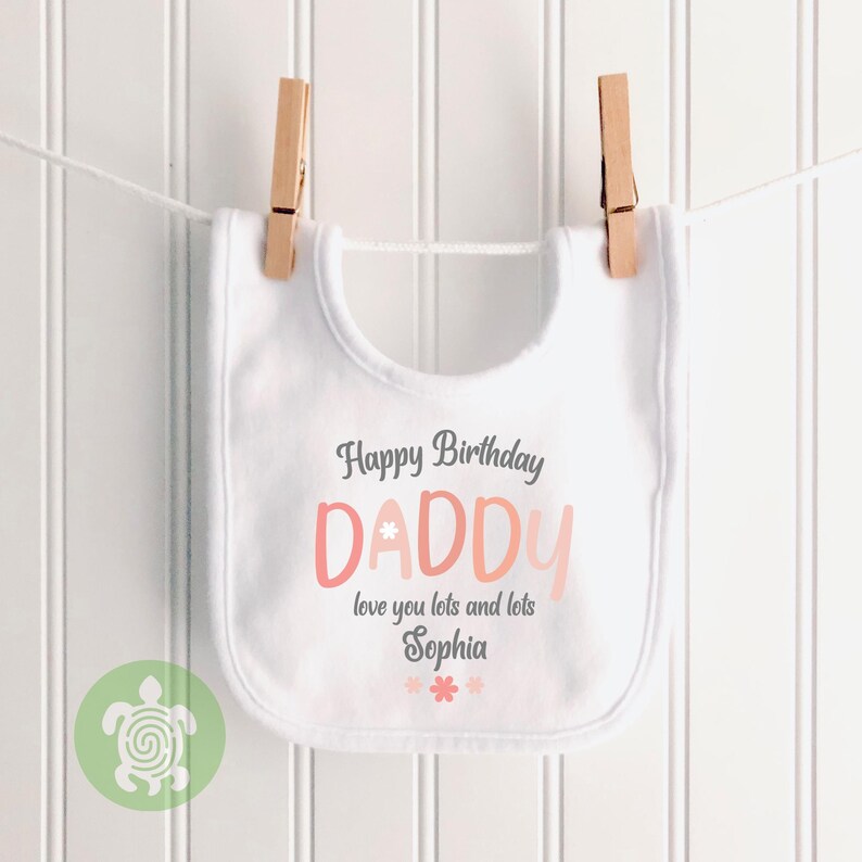 May include: White baby bib with the text "Happy Birthday DADDY" in gray and pink. The bib is hanging from a clothesline with wooden clothespins. The text "love you lots and lots Sophia" is also on the bib.