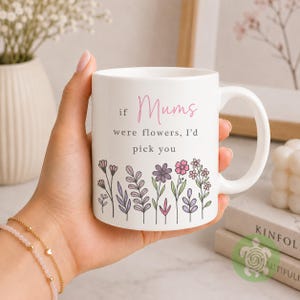 May include: A white ceramic mug featuring the black and pink text 'if Mums were flowers, I'd pick you' printed on the front. Below the text is a colourful illustration of various wildflowers in shades of purple, pink, and green.