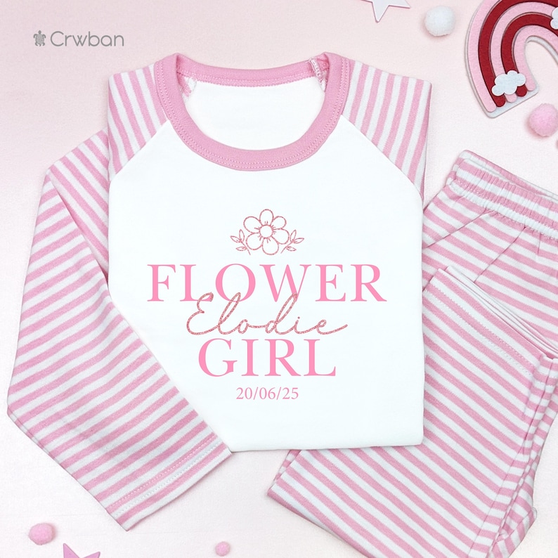 May include: A pink and white striped pyjama set. The top has a white body with pink sleeves and collar. The text "FLOWER Elodie GIRL" is printed in pink, along with a flower graphic and the date 20/06/25.