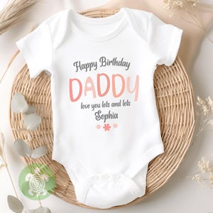 May include: White baby onesie with the text "Happy Birthday DADDY" in gray and pink lettering. The text "love you lots and lots Sophia" is below the word "DADDY", with pink flower graphics at the bottom. The onesie is on a woven basket.