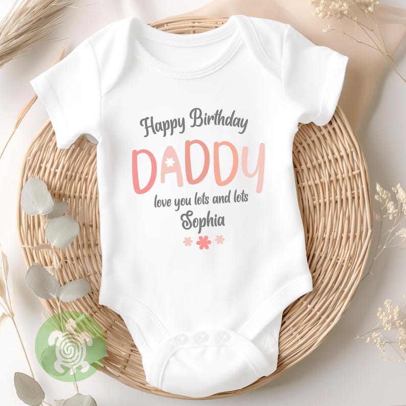 May include: White baby onesie with the text "Happy Birthday DADDY" in gray and pink lettering. The text "love you lots and lots Sophia" is below the word "DADDY", with pink flower graphics at the bottom. The onesie is on a woven basket.